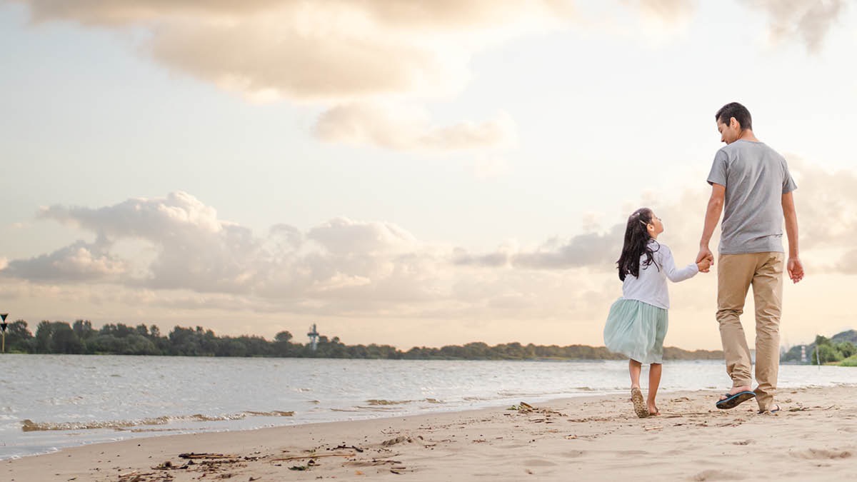 Un padre camina por la playa con su hija y la lleva de la mano.