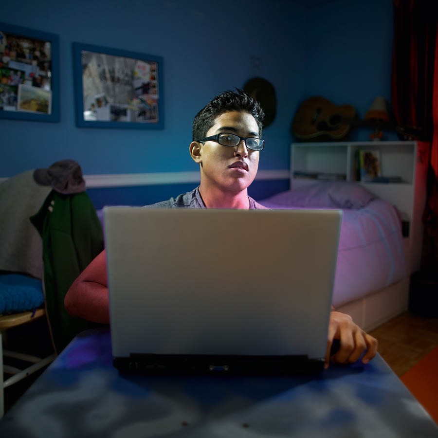 ’n Jong man, alleen in sy kamer, kyk weg van sy rekenaar sodat hy nie na iets kan kyk wat uit die bloute op sy skerm verskyn het nie.