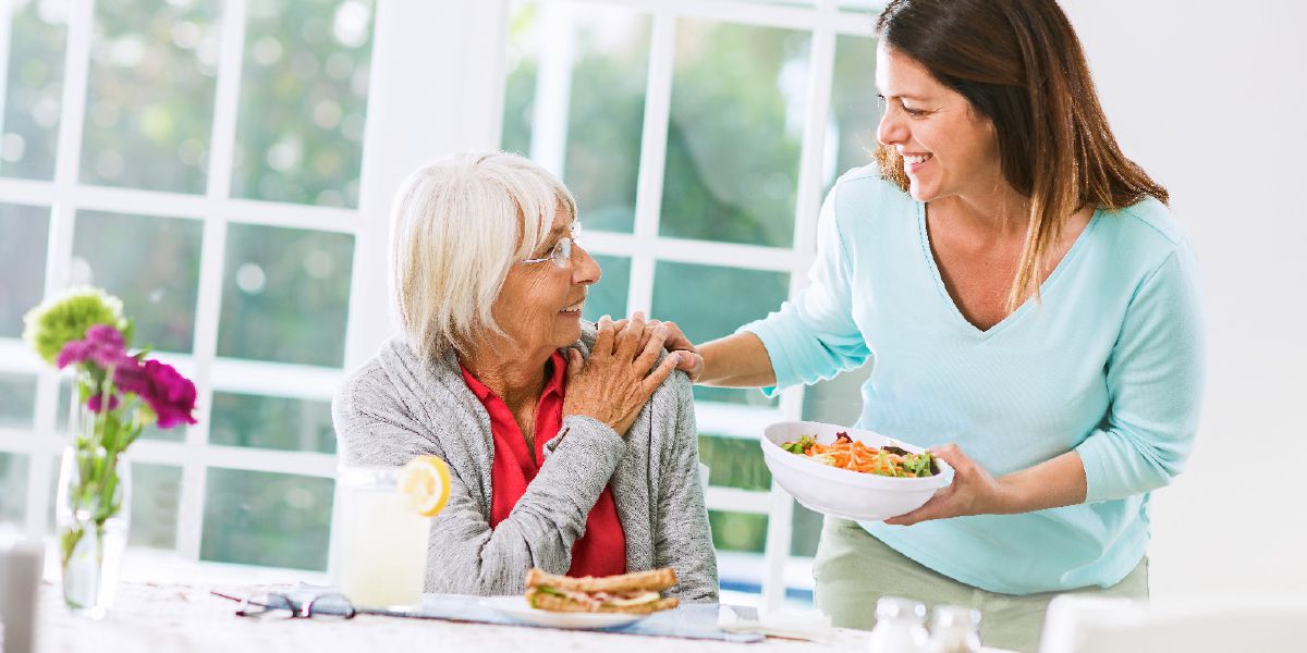 An adult daughter lovingly brings food to her elderly mother