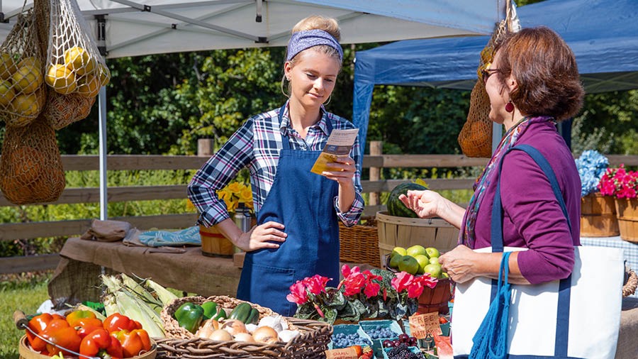 A sister shopping at an outdoor market, giving a tract to a young woman at a vegetable stand.