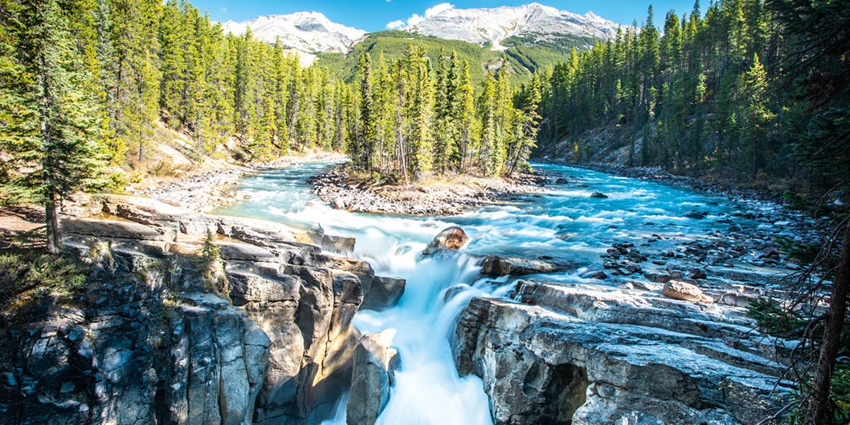 A river flowing toward a waterfall. The river is surrounded by snowcapped mountains and a forest..