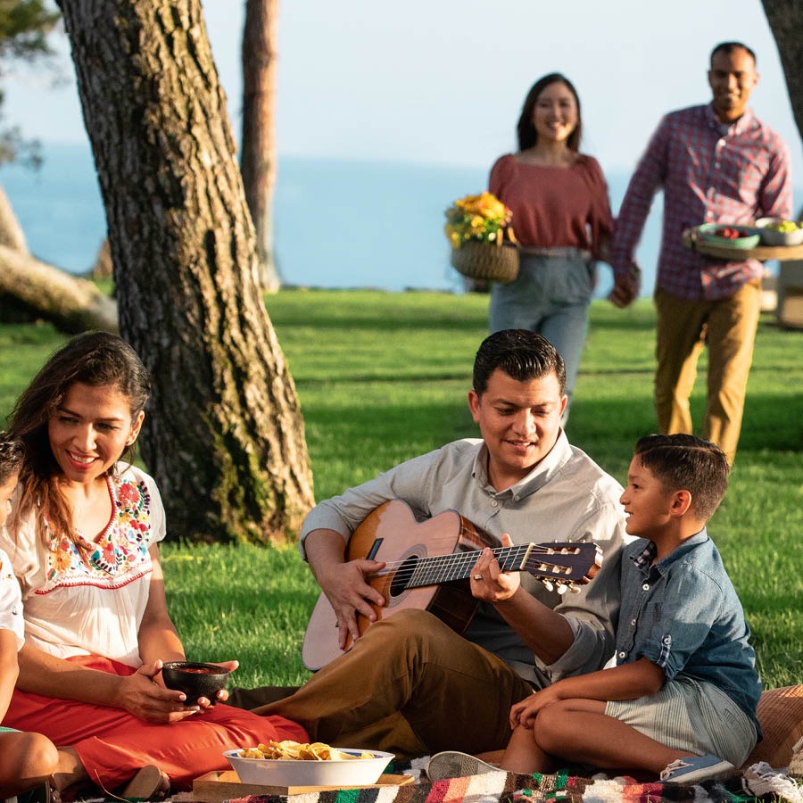 A family enjoying a picnic in a park. The father is playing a guitar.