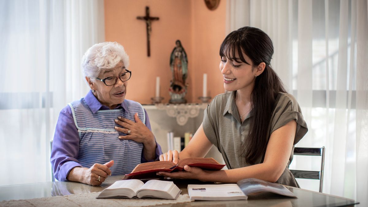 Un hermana le da clases de la Biblia a una mujer mayor, que tiene una cruz en la pared y una imagen en la mesa. A la mujer le impresiona ver el nombre de Dios en su propia Biblia.