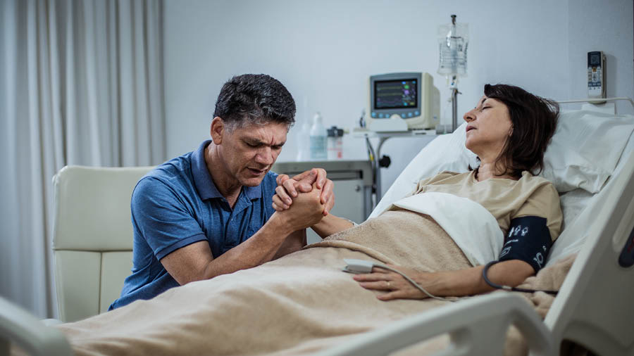 En mand der beder sammen med sin kone, som ligger i en hospitalsseng.
