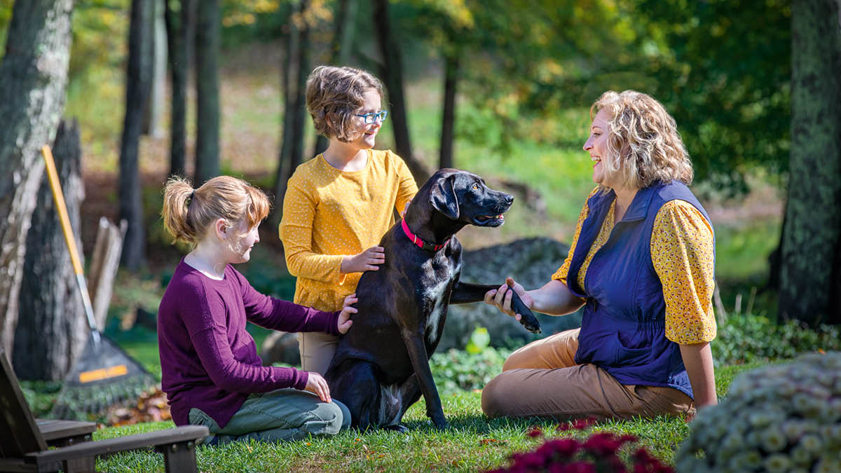Eine Mutter und ihre zwei Töchter sitzen auf dem Gras und streicheln ihren Hund.
