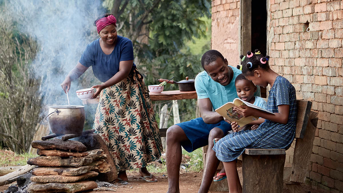 Eine glückliche Familie vor ihrem bescheidenen Zuhause. Die Mutter kocht auf einer Feuerstelle ein einfaches Essen. Ganz in der Nähe liest der Vater mit den beiden Kindern in dem Buch „Was wir aus der Bibel lernen können“.