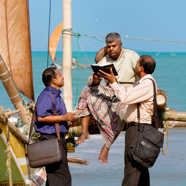 Dos testigos de Jehová predicando a un marinero en la orilla del mar. Le leen un texto de la Biblia