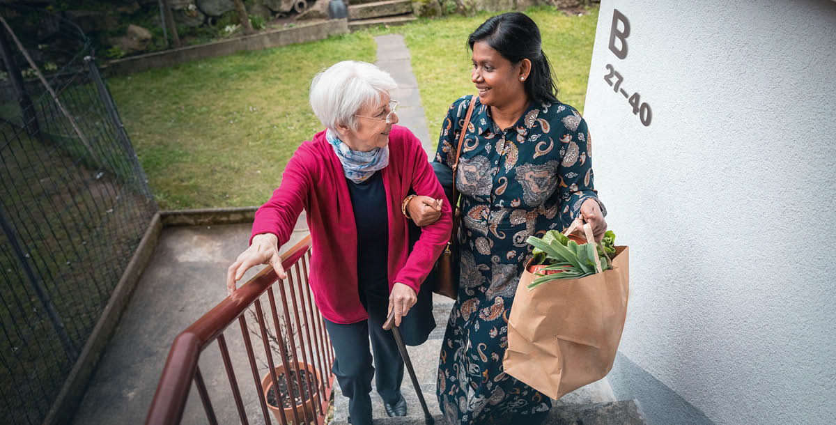 An Indian woman helping an older Caucasian woman up the stairs with her groceries.