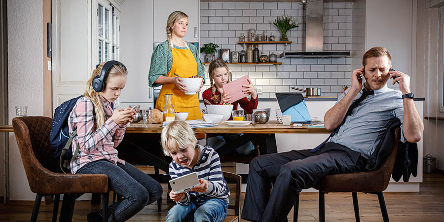 A mother setting breakfast on a table and looking irritated as her husband and children are distracted with their electronic devices.