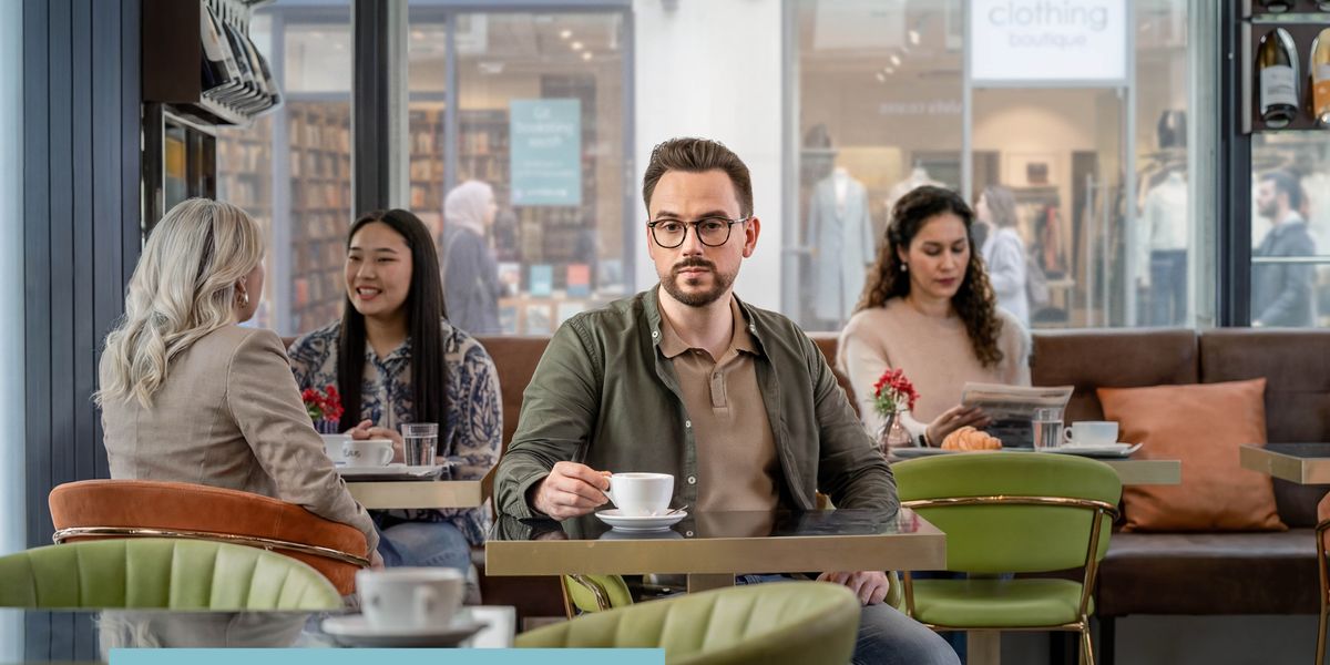 A young man sits alone in a café with a cup of coffee in front of him, gazing into the distance while others read and socialize.