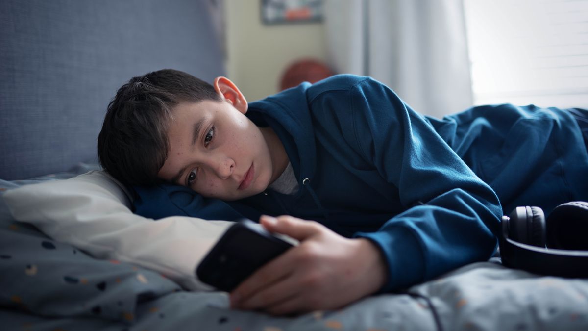 A teenage boy lies on his bed, looking at his phone.