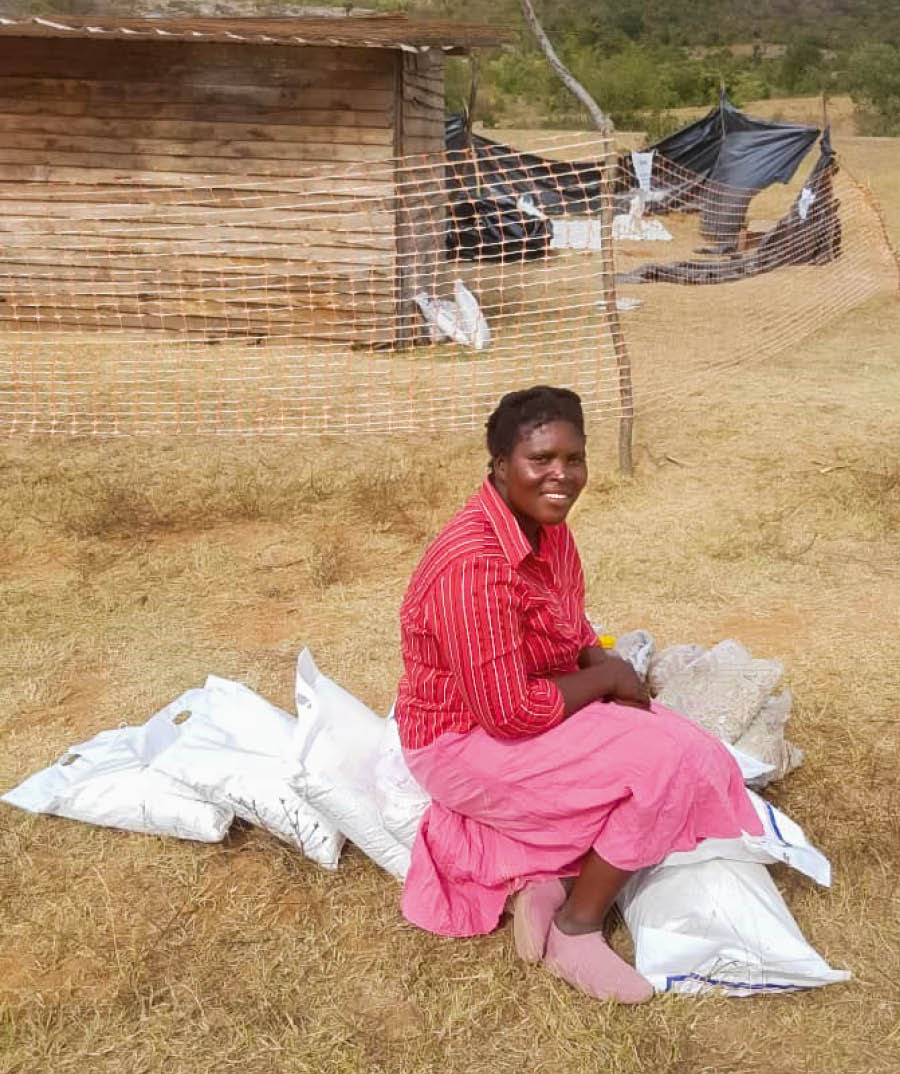 Prisca outside her home, smiling with several bags of food supplies.