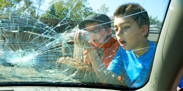 Two young boys looking at a car’s windshield broken by their baseball