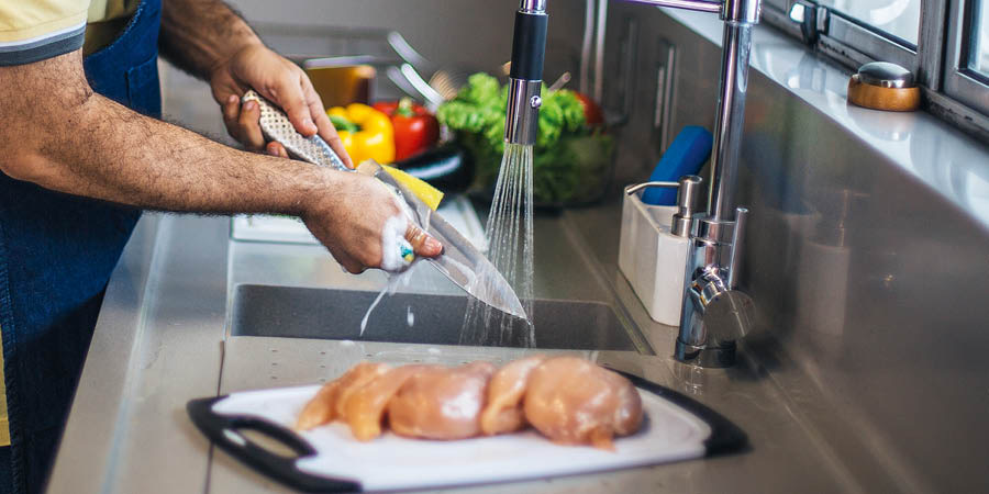 A man washing a knife with soap and water. Raw chicken and fresh vegetables sit on separate cutting boards.