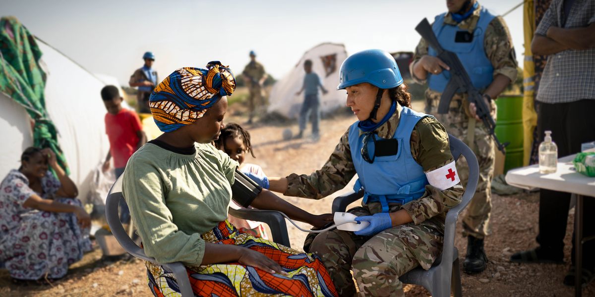 A female medic checking a refugee woman’s vital signs. Other refugees and armed peacekeepers are nearby.