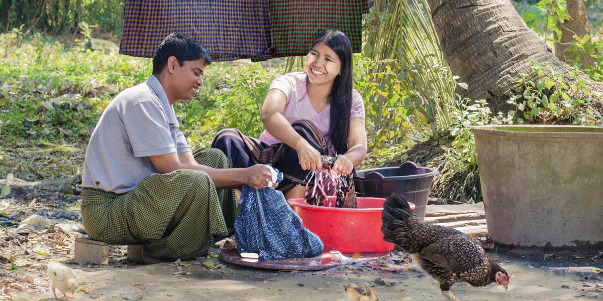 A husband sitting on the ground next to his wife and happily washing clothes with her.