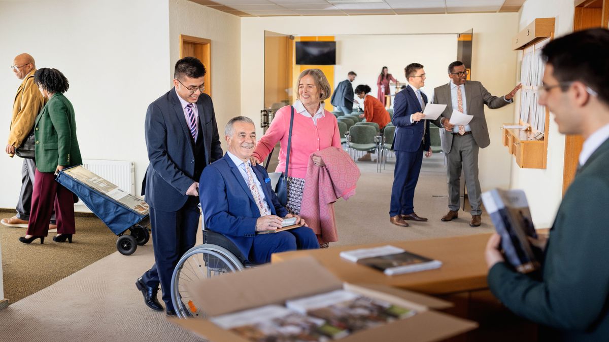 A brother helps an elderly brother in a wheelchair at a Kingdom Hall. Others nearby clean the auditorium and tend to other congregation tasks, including organizing literature and updating the information board.