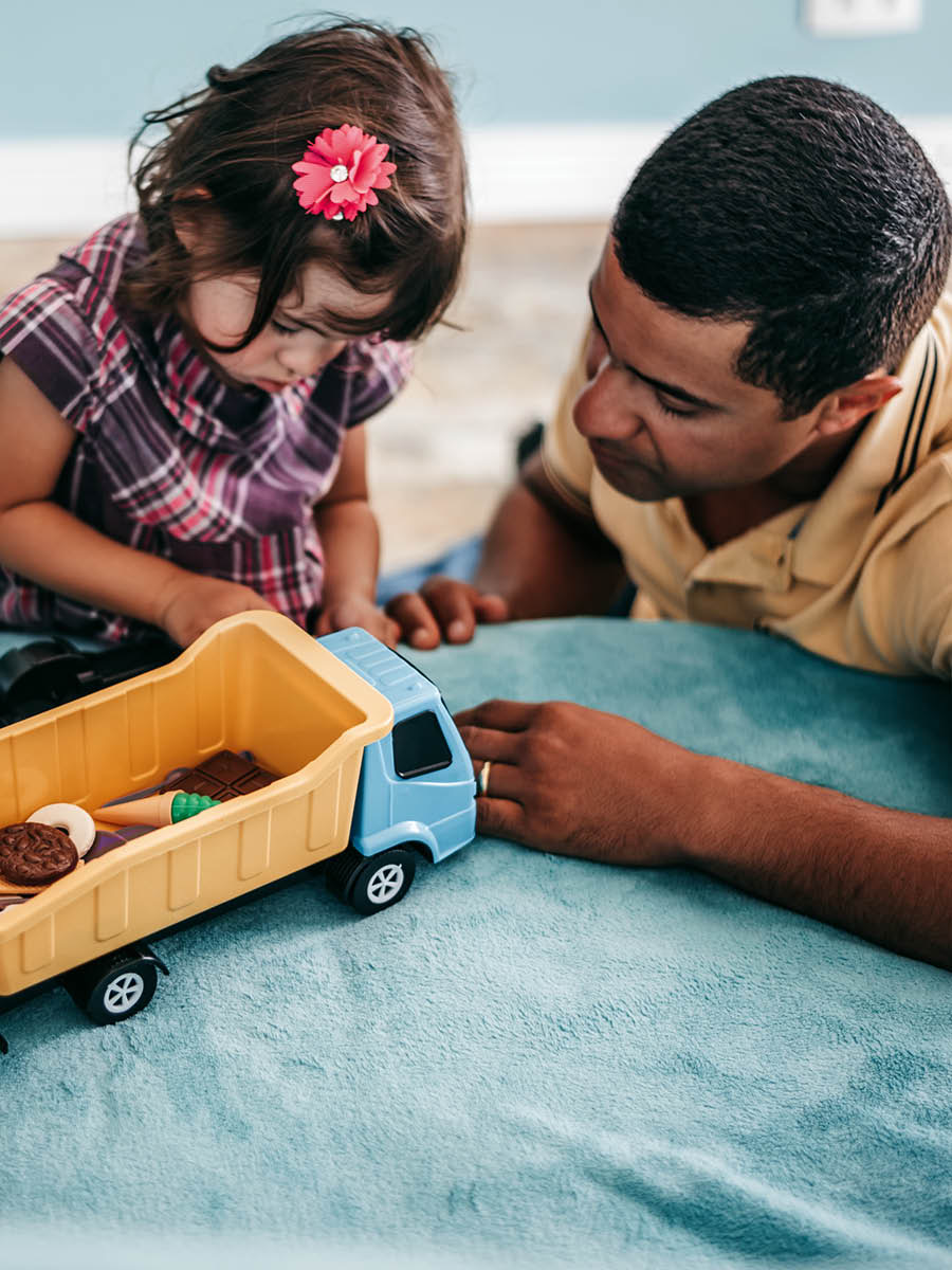 A father sitting next to his young daughter as she plays with some toys.