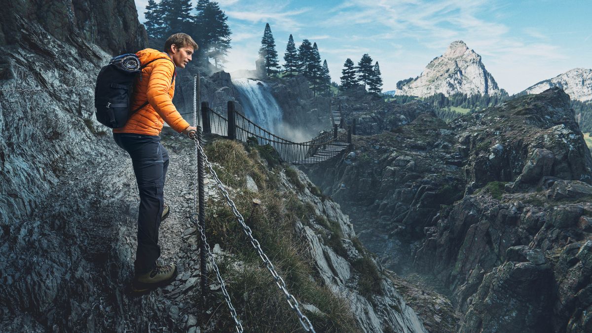 The young man shown in previous articles walking on a steep and narrow path that has a chain railing. A suspension bridge and a mountain peak can be seen in the distance.