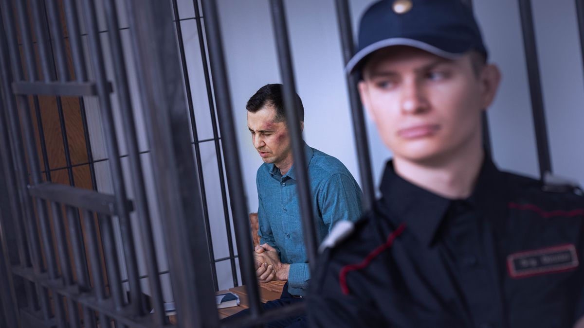 Un hermano con moretones en la cara ora en su celda de prisión mientras un guardia lo custodia.