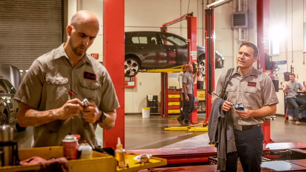 Un hermano, que trabaja en un taller de reparación de vehículos, acercándose a un compañero de aspecto intimidante para darle una invitación a las reuniones. El resto de compañeros de trabajo se están riendo del hermano.
