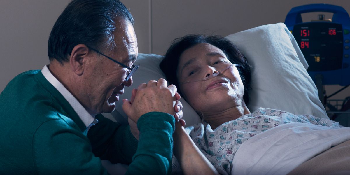 Egy testvérnő kórházi ágyon fekszik; a férje fogja a kezét, és imádkozik vele