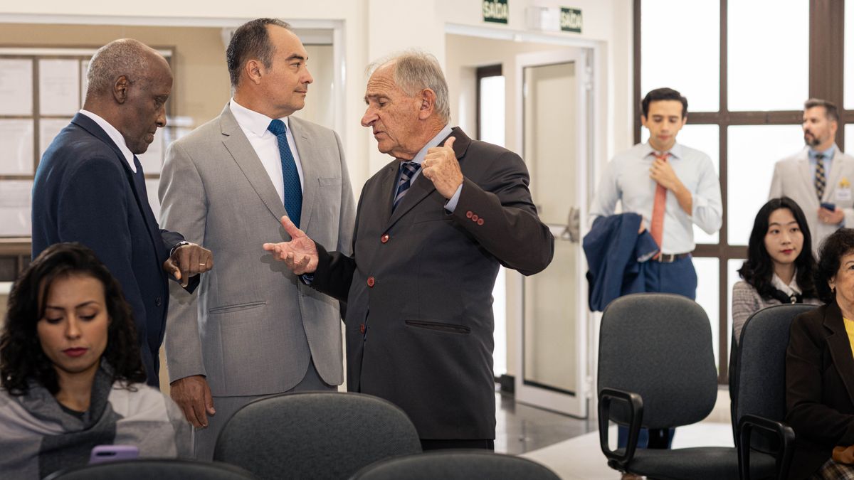 L’anziano ascolta altri due anziani che dicono qualcosa di negativo riguardo al giovane fratello, che sta entrando in tutta fretta nella Sala del Regno mentre sta per iniziare l’adunanza.