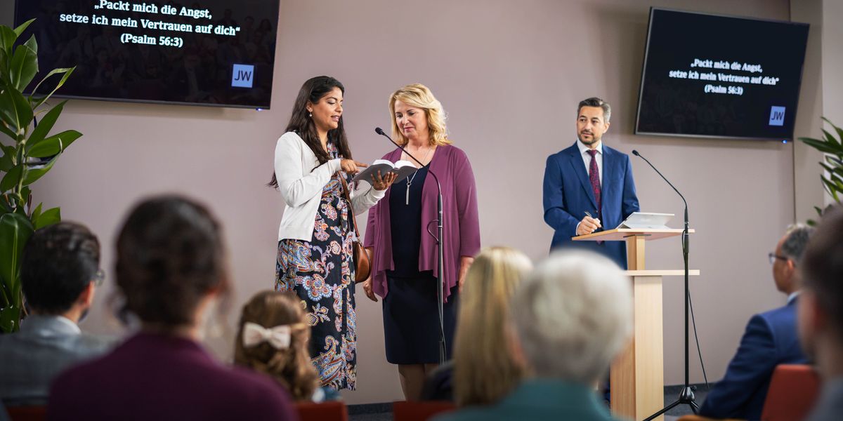 En la plataforma de un Salón del Reino, dos testigos de Jehová escenifican una escena de la predicación en la reunión de entre semana.