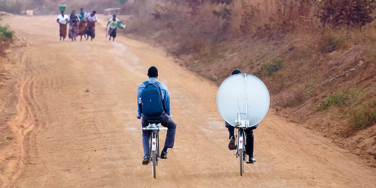 Broeders die met de fiets via een zandweg satellietapparatuur naar hun gemeente vervoeren.