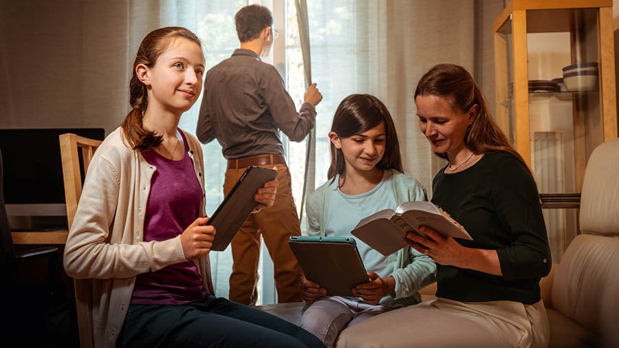 Een broeder kijkt thuis uit het raam terwijl zijn vrouw met hun twee dochters de Bijbel leest.