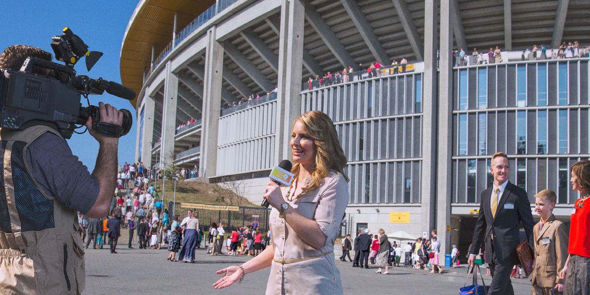 Ekipa telewizyjna przed stadionem, na którym odbywa się kongres Świadków Jehowy; na drugim planie widać odpowiednio ubranych uczestników kongresu