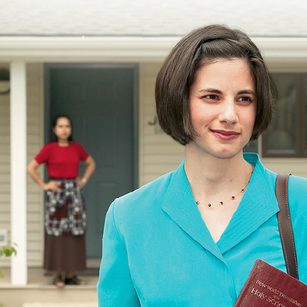 Uma irmã na pregação saindo confiante da frente de uma casa enquanto a moradora irritada está na porta e a observa.