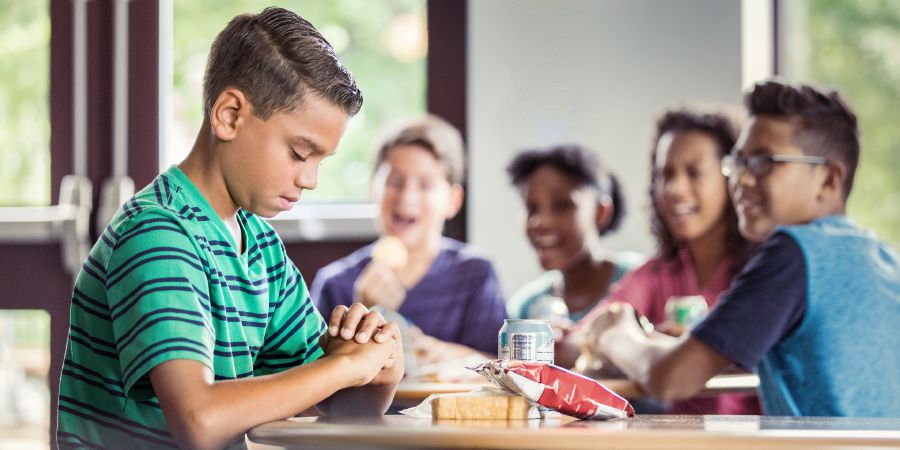 Um menino Testemunha de Jeová faz oração antes de comer e outros meninos riem dele