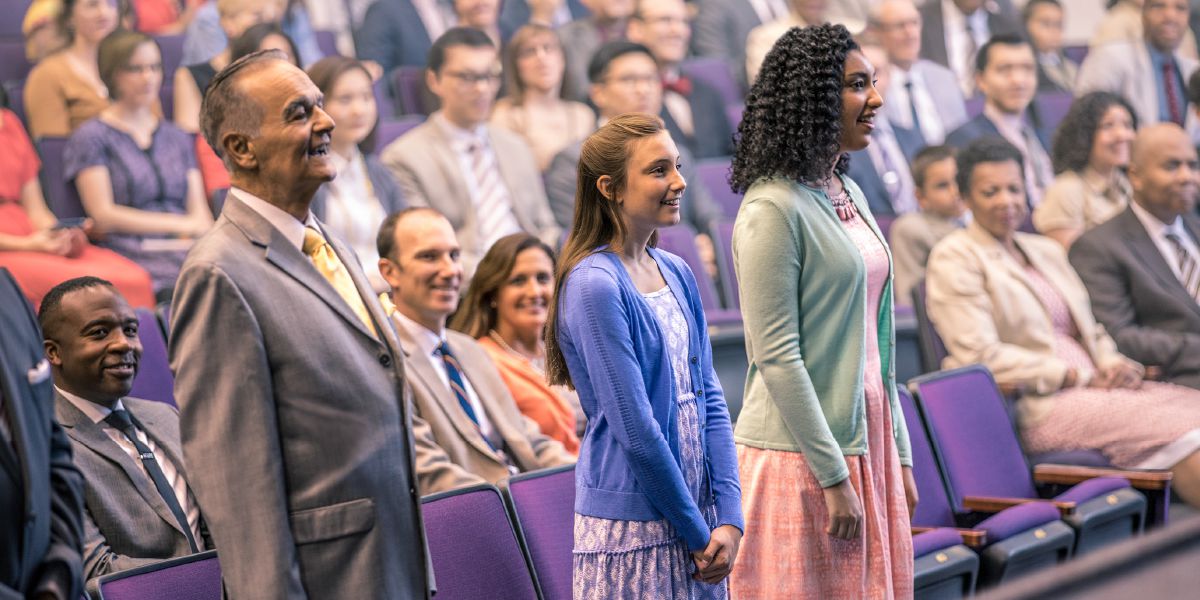 Un señor mayor y dos jovencitas están de pie porque van a responder las preguntas del discurso del bautismo. Detrás los miran satisfechos los padres de una de ellas y el maestro de la Biblia del señor