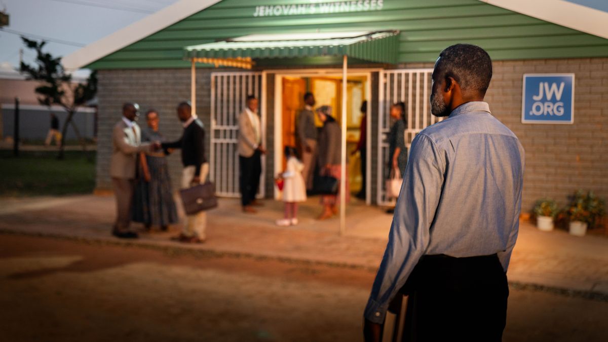 Ntate ea holileng o tšoere Bibele a eme hojana le Holo ea ’Muso. O shebile bara le barali ba bo rona ha ba ntse ba amohelana ka mofuthu ha ba fihla libokeng.