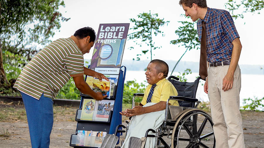 Mark Herman sitting in a wheelchair next to a literature cart, preaching to a man.